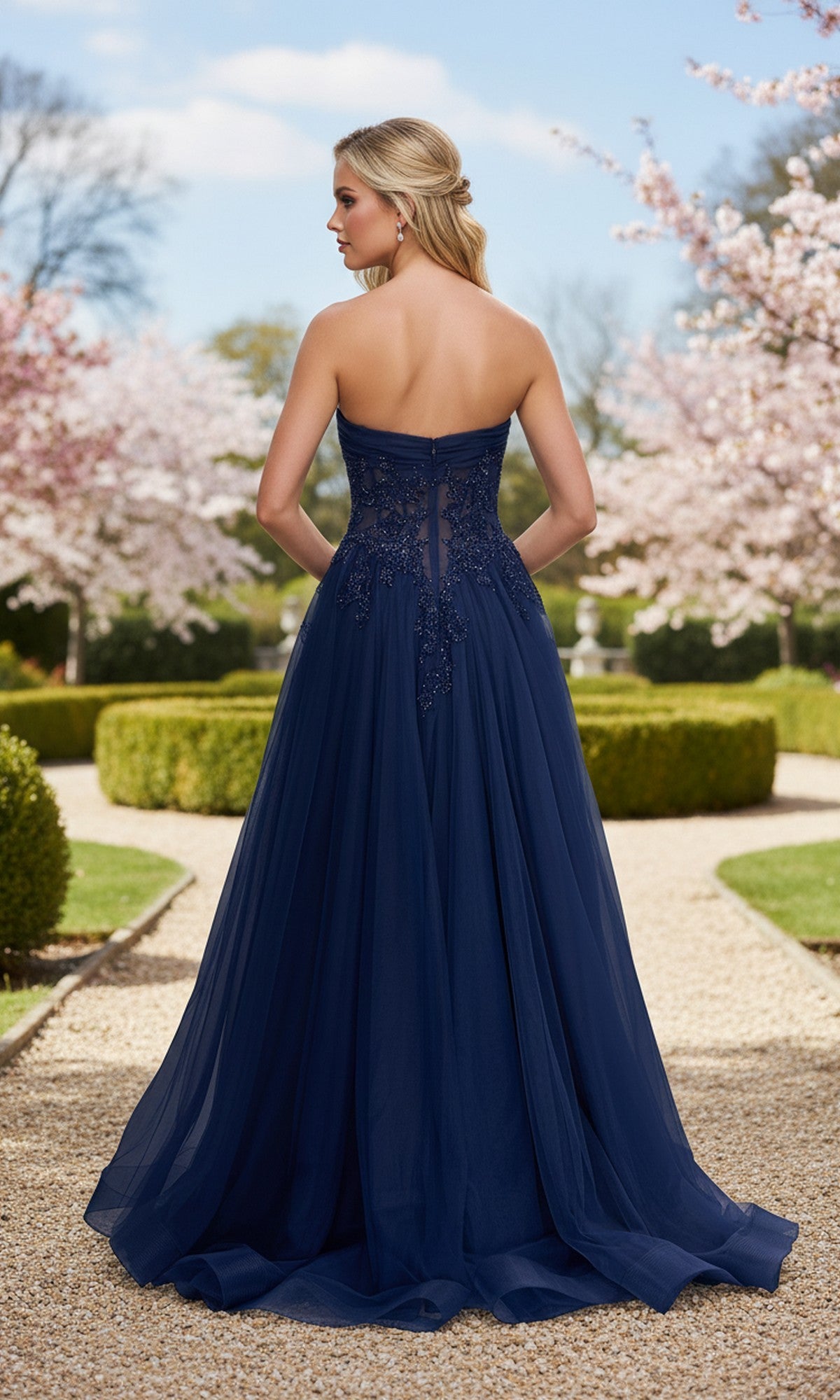 Wearing the La Femme 33528 long prom dress with a strapless, sheer corset bodice, a woman stands outdoors on a gravel path among blooming trees and manicured hedges.